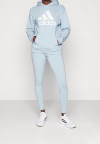 Lichtblauwe hoodie met groot wit Adidas-logo, gecombineerd met lichtblauwe leggings. Geweven stof met een zachte uitstraling, inclusief zakken.