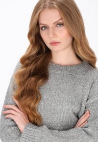 Jasnoszary sweter dzianinowy z okrągłym dekoltem, teksturowaną powierzchnią i długimi rękawami, noszony przez osobę z falowanymi włosami, skrzyżowanymi ramionami.