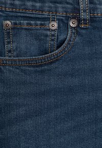 Tmavě modrá džínovina s viditelným šitím, která má malou přední kapsu a kovové nýty. Hladká textura s klasickým designem džínů.