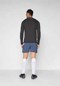 Man som bär svart långärmad tröja, blå atletiska shorts, vita knästrumpor och svarta fotbollsskor, står vänd bort från kameran mot en enhetlig bakgrund.