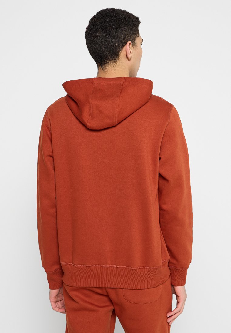 Rdzawoczerwony hoodie z kapturem na sznurki, ściągaczami przy rękawach i na dole. Miękka faktura tkaniny bez widocznych logo czy wzorów.