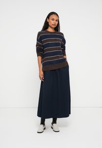 Egy nő áll, barna és sötétkék csíkos pulóvert, hosszú sötétkék szoknyát, fekete zoknit és fehér slip-on cipőt visel egy fehér háttér előtt.