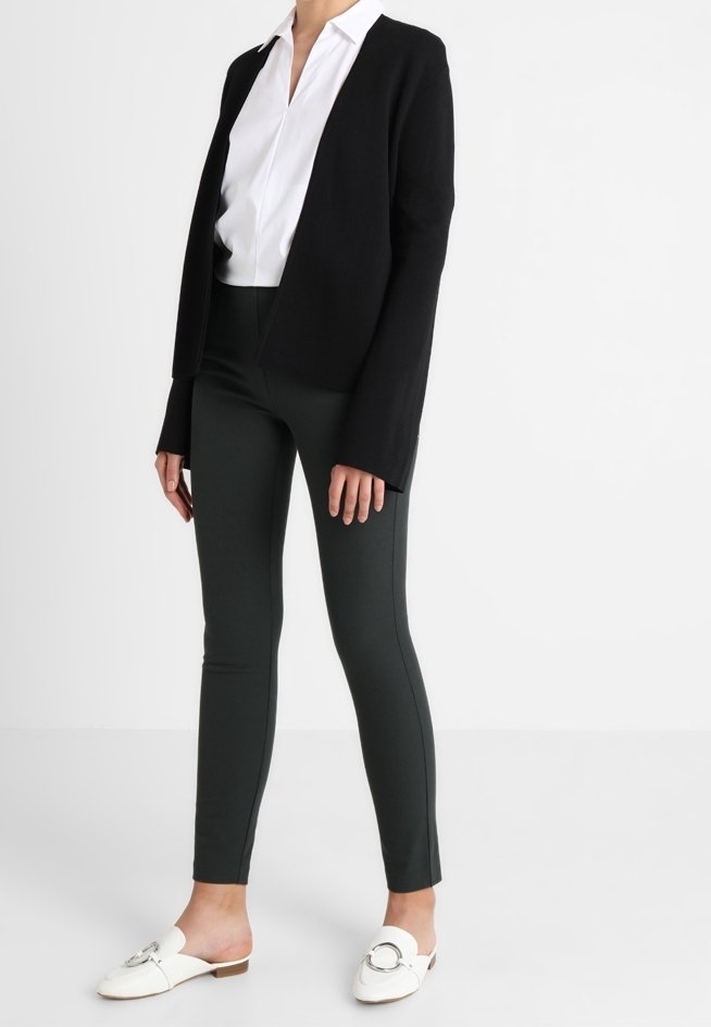 Black knit cardigan, white collared shirt, dark green slim-fit pants, white slip-on shoes with a circular accent, smooth textures throughout.