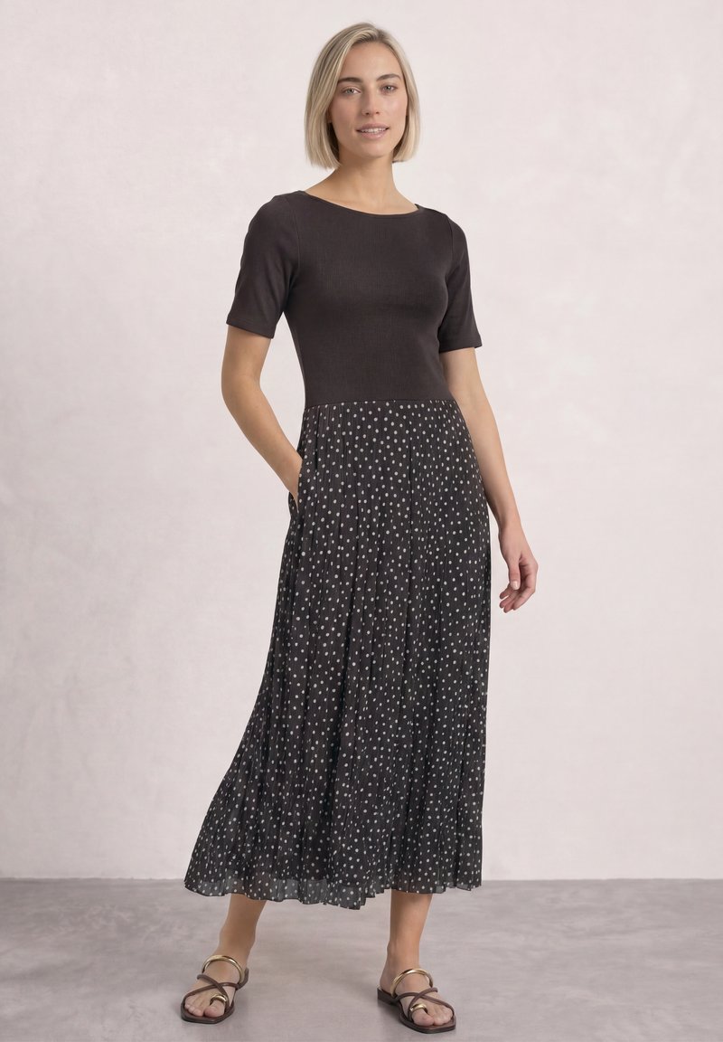 Femme aux cheveux blonds courts portant un haut noir à manches courtes et une jupe plissée noire à pois blancs, debout avec une main dans la poche.