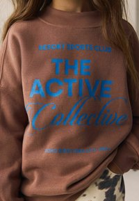 Hnedá mikina s okrúhlym výstrihom a výrazným modrým nápisom „RESORT SPORTS CLUB“ a „THE ACTIVE Collective“. Mäkká textúra.