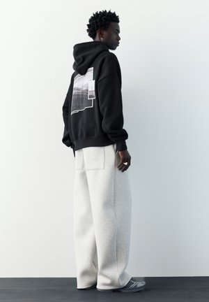 Jonge man staand zijwaarts, gekleed in een zwarte hoodie met grafische print op de achterkant, losse lichtgrijze broek en grijze sneakers, op een donkere vloer.