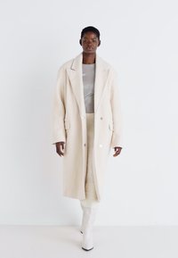 Manteau en laine de couleur crème avec des revers surdimensionnés, de grands boutons et des poches latérales, associé à un haut gris clair et une jupe texturée couleur crème.