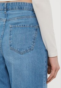 Ανοιχτό μπλε τζιν παντελόνι με ψηλή μέση, που διαθέτει μια μοναδική πίσω τσέπη και ελαφριά φθορά. Η υφή είναι λεία με ορατές ραφές.