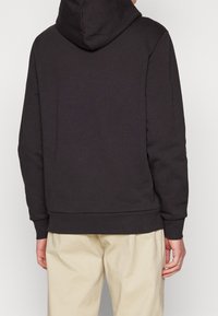 Hoodie gris foncé avec une poche kangourou, des poignets et un ourlet côtelés, doté d'une capuche avec cordon de serrage et d'une texture de tissu douce. Porté avec un pantalon de couleur claire.