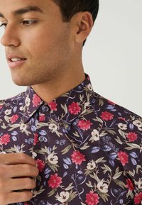 Korte mouwen bloemenoverhemd in donkerpaars, met roze en witte bloemen, groene bladeren en blauwe accenten. Kenmerken zijn een button-down kraag.