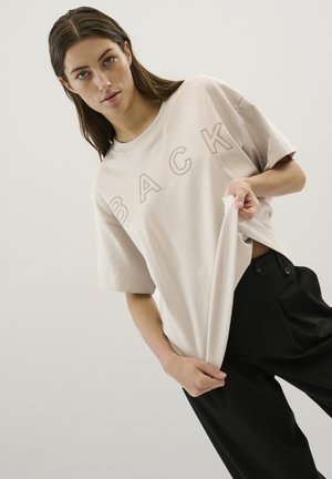 T-shirt en coton beige clair avec "BACK" imprimé en une teinte plus foncée. Coupe décontractée, manches courtes et col rond. Associé à un pantalon noir.