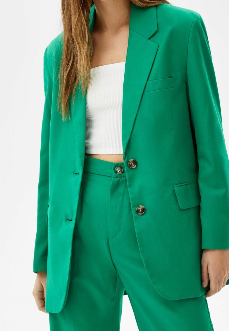 Blazer vert avec un devant boutonné, une poche poitrine simple, et des revers crantés, associé à un pantalon vert assorti. Texture de tissu lisse.