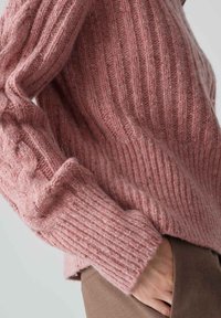 Personne portant un pull en tricot côtelé rose, une main glissée dans la poche d'un pantalon marron, sur un fond clair.