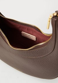 Borsa in pelle marrone con texture e zip dorata. All'interno presenta una fodera bordeaux e un piccolo etichetta incisa con "Coccinelle."