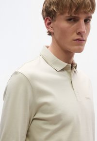 Ανοιχτό μπεζ πόλο μπλουζάκι από λείο, μαλακό ύφασμα· διαθέτει γιακά και διακριτικό λογότυπο στο στήθος, με εφαρμοστή σιλουέτα.