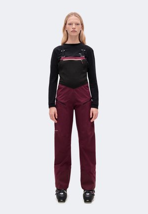 Un mannequin se tient de face, vêtu d’un pantalon de ski bordeaux à bretelles noires, d’un haut noir et de chaussures de ski, sur un fond uni.