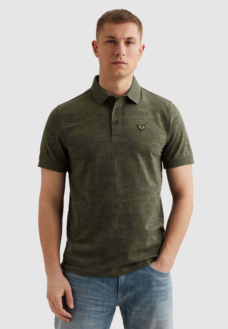 Jeune homme portant un polo vert olive texturé avec un petit emblème brodé jaune et noir, associé à un jean bleu clair.