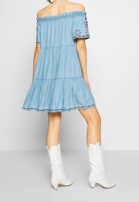 Nainen, joka pukeutuu vaaleansiniseen olkaimettomaan mekkoon, jossa on kirjailuja hihoissa, ja valkoisiin polvimittaisiin korkokengät, seisoo tasaisen taustan edessä.