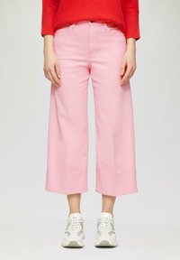 Femme portant un pantalon large cropped taille haute rose, des baskets blanches et un pull côtelé rouge se tenant devant un fond uni.