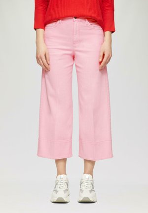 Vrouw in hooggewaisted roze wijde cropped broek, witte sneakers en een rode geribbelde trui staat tegen een effen achtergrond.