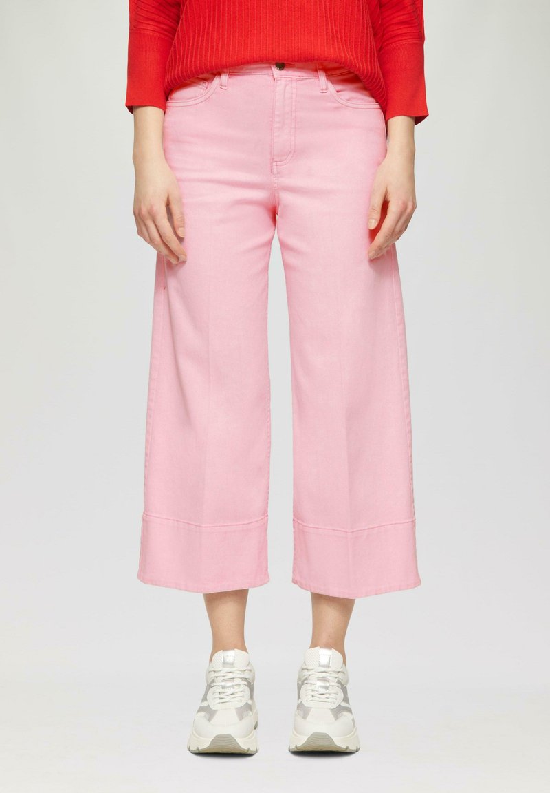 Femme portant un pantalon large cropped taille haute rose, des baskets blanches et un pull côtelé rouge se tenant devant un fond uni.