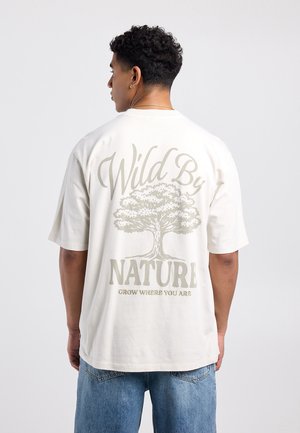 Man draagt wit T-shirt met boomgrafiek en tekst "Wild By Nature, Grow Where You Are" en staat tegen een effen achtergrond.