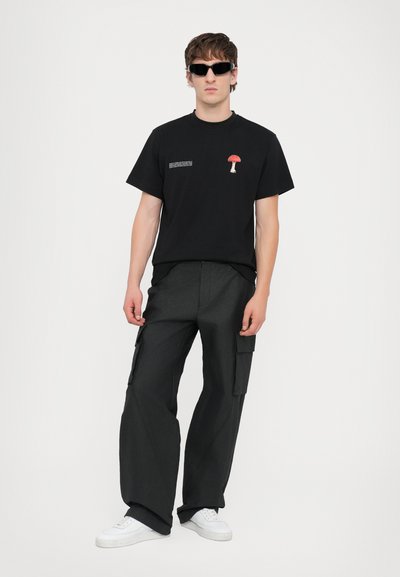 T-shirt noir avec un graphique de champignon rouge et du texte sur le côté gauche, associé à un pantalon cargo sombre et des baskets blanches.