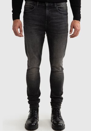 Mann trägt schwarze, ausgewaschene Slim-Fit-Jeans und schwarze Leder-Schnürstiefel, steht vor einem schlichten Hintergrund.