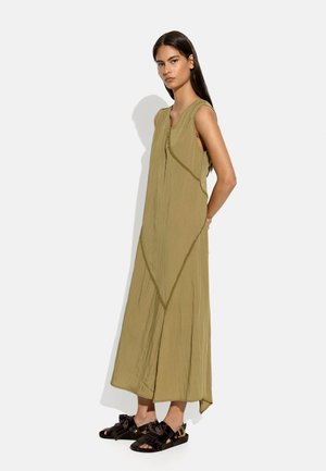 Kobieta stojąca w bezrękawnikowej oliwkowozielonej sukience midi z ukośnymi szwami, w połączeniu z czarnymi sandałami z frędzlami i odkrytymi palcami na białym tle.