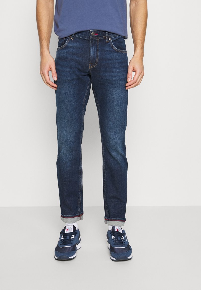 Dunkelblaue Jeans im geraden Schnitt mit sichtbaren Nähten und aufgerolltem Saum. Kombiniert mit marineblauen Turnschuhen.