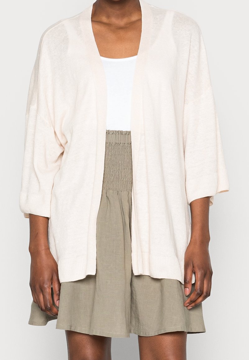 Cardigan rose pâle à ouverture devant, manches larges, poignets côtelés et texture douce, porté sur un haut blanc et une jupe kaki.