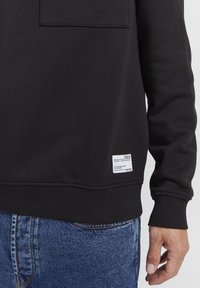 Sort sweatshirt med en frontlomme, ribbede manchetter og kant. Inkluderer et mærke med tekst i nederste højre hjørne. Kombineret med denim.