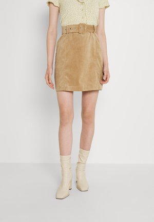 Person iført beige ruskind mini nederdel med bredt bælte, gul blomstret kortærmet bluse og cremefarvede ankelstøvler med hæl, stående mod en hvid baggrund.