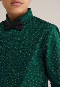 Zelena košulja s gumbima koja sadrži geometrijski uzorak s malim bijelim kvadratima, uparen s crnim leptir-mašnom. Glatka tkanina i strukturirani ovratnik.