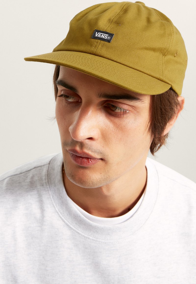 Grön keps med rak brätte, tillverkad av bomull, med en svart Vans-logotyp på framsidan. Bärs med en ljusgrå crewneck-tröja.