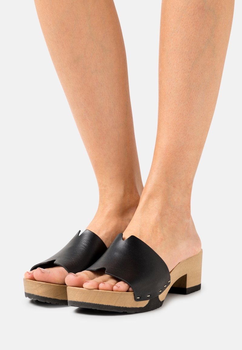 Pieds portant des sandales en cuir noir à bout ouvert avec talons blocs en bois sur un fond clair.