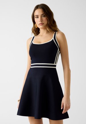 Kobieta w bezrękawnikowej czarnej sukience z białą lamówką i rozkloszowaną spódnicą, stojąca na tle jednolitego jasnego tła.