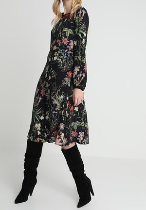 Robe florale en tissu noir avec un motif botanique coloré, à manches longues et jupe évasée. Associée à des bottes noires jusqu'aux genoux.