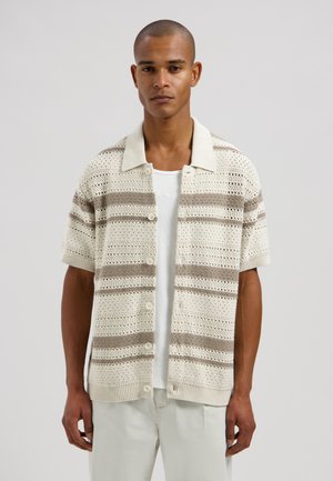 Man draagt een korte mouwen, gebreid vest met knopen in crème- en beige strepen over een wit overhemd met lichte broek, staand tegen een effen achtergrond.