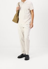 Polo beige clair avec motif texturé, pantalon blanc léger et chaussures noires. Le modèle porte un sac fourre-tout marron.