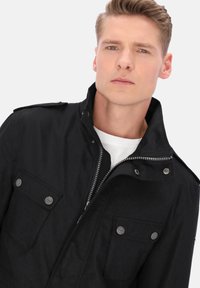 Sort lynlåstrøje med høj krave, to brystlommer, sølvfarvet hardware og et diskret teksturmønster, kombineret med en hvid skjorte under.