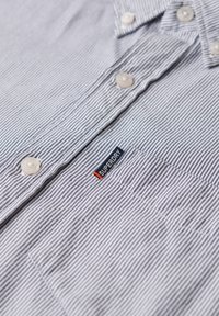 Camicia button-up con motivo a righe blu e bianche, bottoni bianchi e una piccola etichetta Superdry blu e arancione sul taschino.