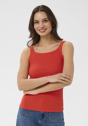 Smilende kvinde med langt mørkt hår iført en rød ærmeløs top og blå jeans, med armene krydset foran en ensfarvet baggrund.