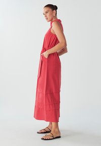 Červené bezrukávové maxi šaty s vrstveným spodným okrajom, viazaným pásom a bočnými vreckami; doplnené čiernymi plochými sandálmi na svetlom pozadí.