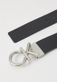 Cinturón de cuero negro con un diseño texturizado, hebilla de metal plateado con un logo estilizado y un cierre rectangular para abrochar.
