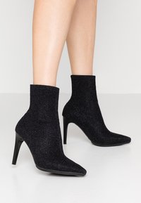 Botas negras hasta el tobillo hechas de un tejido brillante, con punta afilada y tacón alto de aguja. Textura suave con un ajuste ceñido alrededor del tobillo.