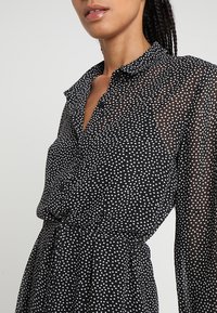 Abito nero trasparente con bottoni, decorato con pois bianchi, maniche lunghe e vita arricciata. Collo con dettagli di texture sottili.