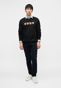 Μαύρο φούτερ με τέσσερα πολύχρωμα γραφικά μπαλώματα στο μπροστινό μέρος, σε συνδυασμό με γαλάζιο πουκάμισο με γιακά, σκούρο τζιν και λευκά αθλητικά παπούτσια.