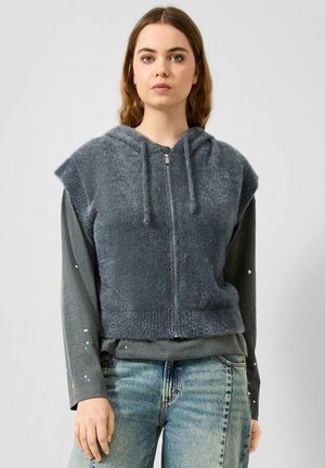 Jeune femme portant un sweat à capuche gris duveteux sans manches avec fermeture éclair sur un t-shirt à manches longues foncé avec des détails en perles et un jean bleu clair.