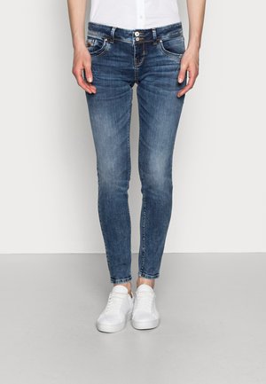 Osoba ubrana w dopasowane niebieskie dżinsy i białe trampki, stojąca na tle jednolitego jasnego tła.